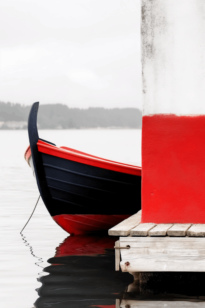 A red and black boat docked by a red and white pier, reflecting on calm water with a blurred forest backdrop