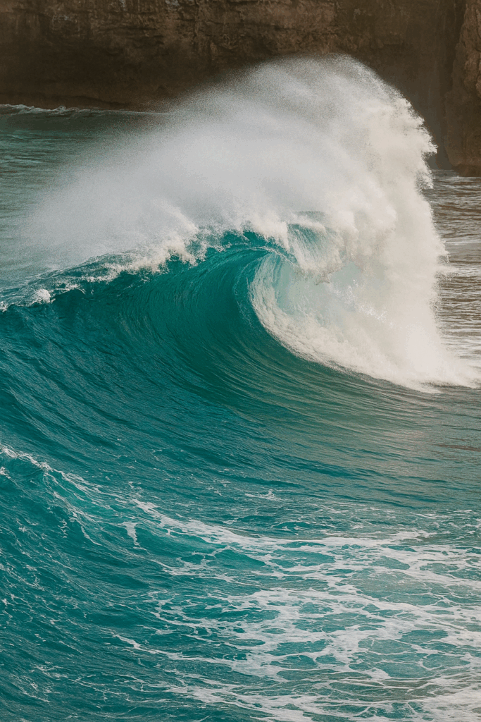 A powerful turquoise ocean wave curling forward as white spray bursts upward, with rocky cliffs faintly visible in the background.
