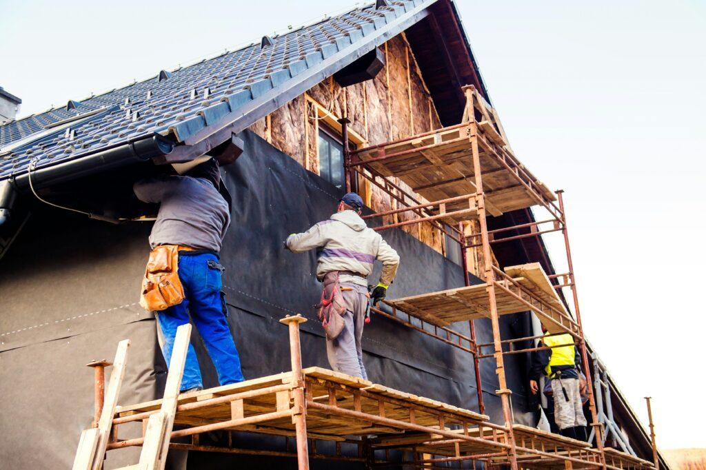 Three construction workers standing on metal scaffolding while installing exterior sheathing and weatherproofing on a residential house, with exposed roof tiles, unfinished wooden framing, and black protective building wrap covering the walls during renovation work