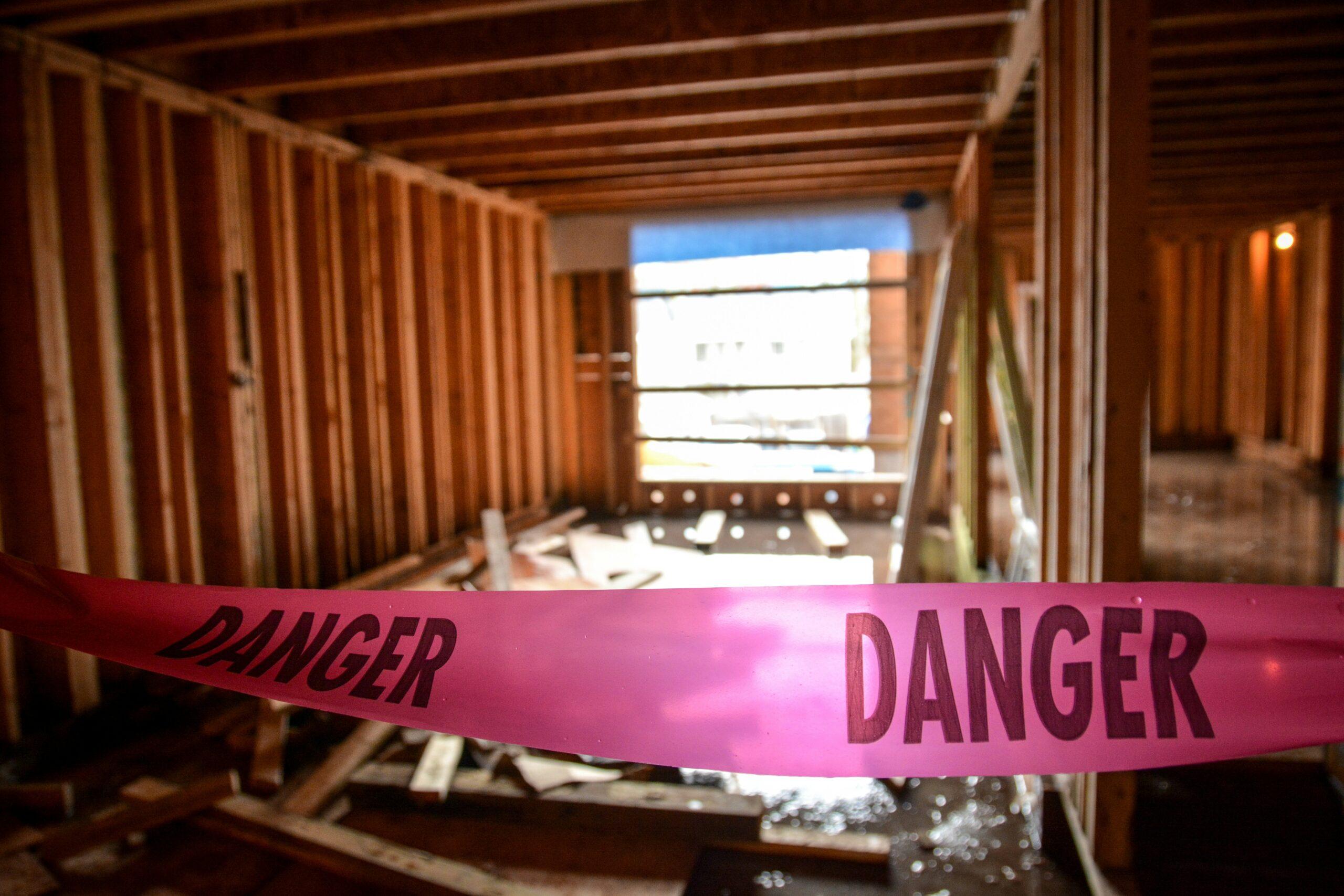Pink “Danger” caution tape stretched across an unfinished interior construction space with exposed wooden framing, scattered debris on the floor, and a partially visible window letting in daylight in the background