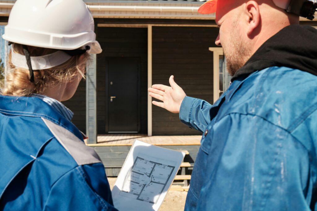 Two construction workers wearing hard hats standing in front of a house, reviewing floor plans while one gestures toward the building.