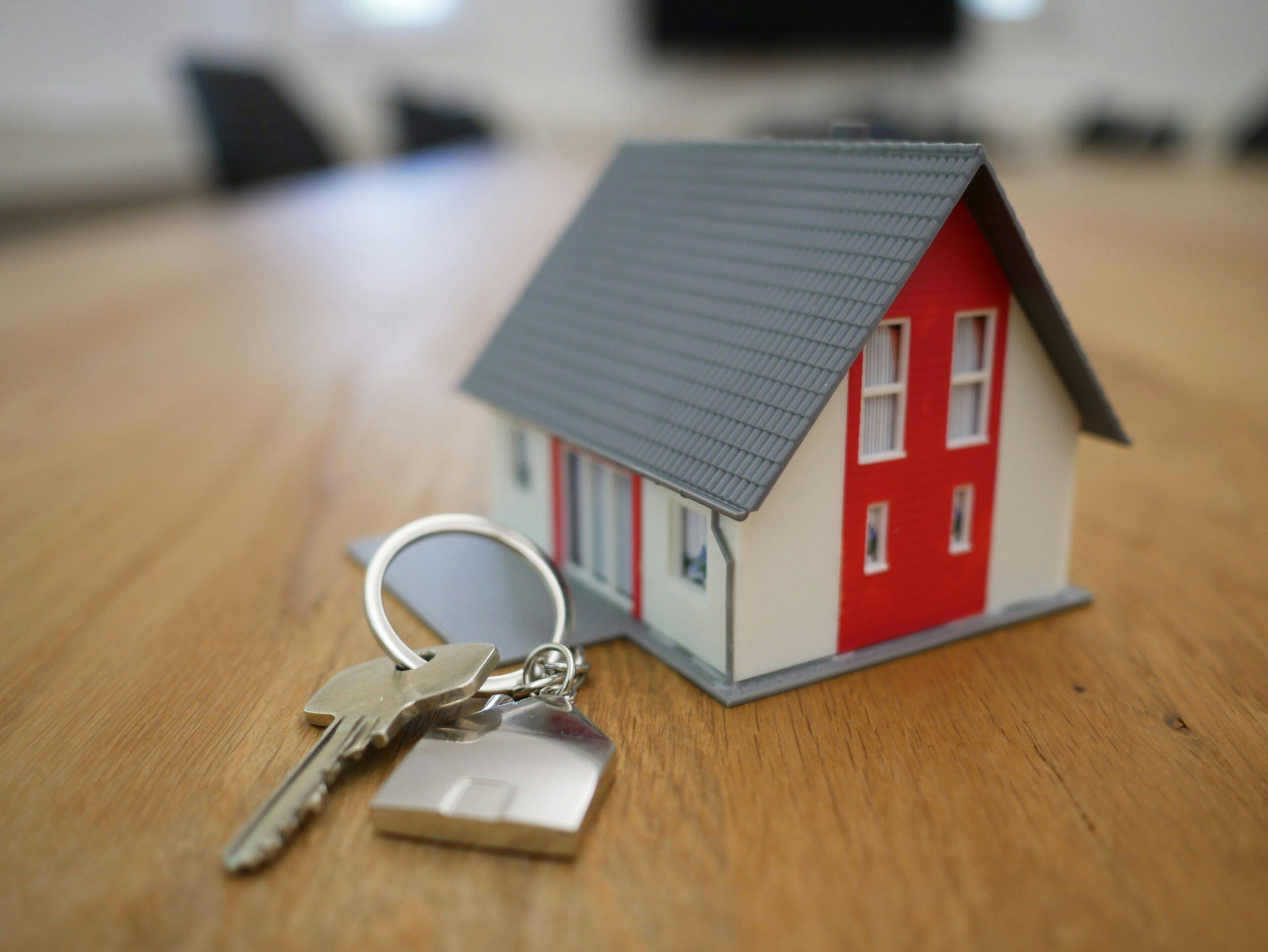 A small house model placed on a wooden table beside a set of house keys with a metal house-shaped keychain.