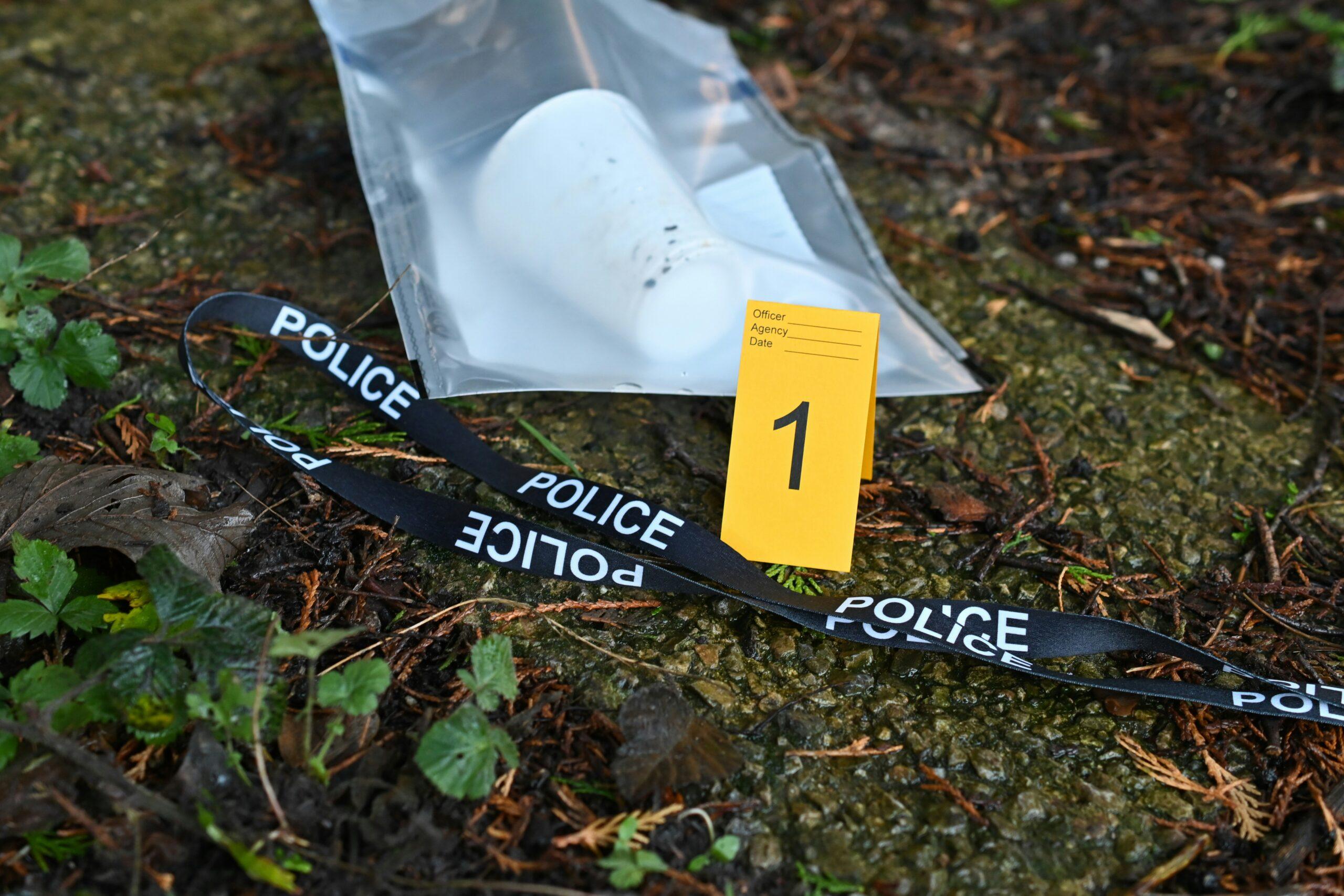 A close-up view of a crime scene on damp ground, showing a sealed transparent evidence bag containing a white disposable cup, a black police tape marked “POLICE” lying across the surface, and a yellow numbered evidence marker labeled “1,” surrounded by leaves, dirt, and small plants in an outdoor setting.
