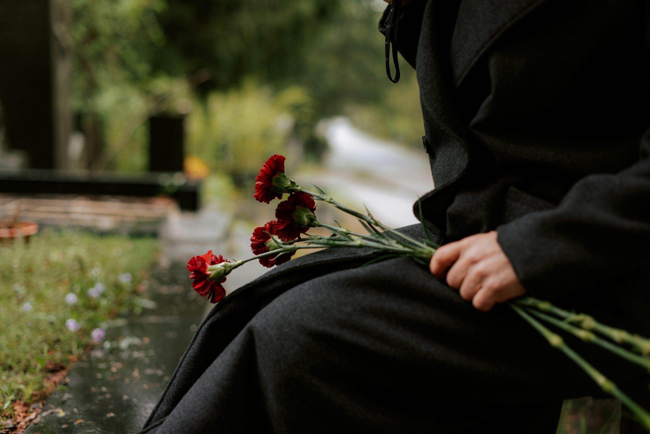 A person dressed in dark clothing sitting outdoors on a bench, holding several red carnations in one hand, with a blurred pathway, greenery, and a somber, quiet atmosphere in the background.