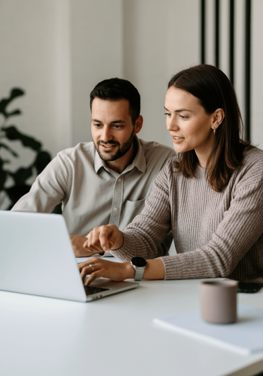 Two colleagues collaborating on a laptop at a desk, with a casual and professional vibe. A mug and notebook add to the workspace setting.