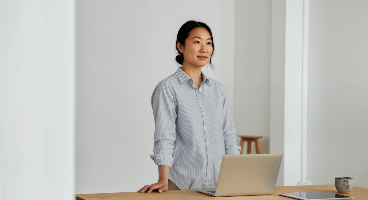 A woman in a light blue shirt stands at a desk with a laptop, mug, and tablet, in a minimalist, well-lit room.