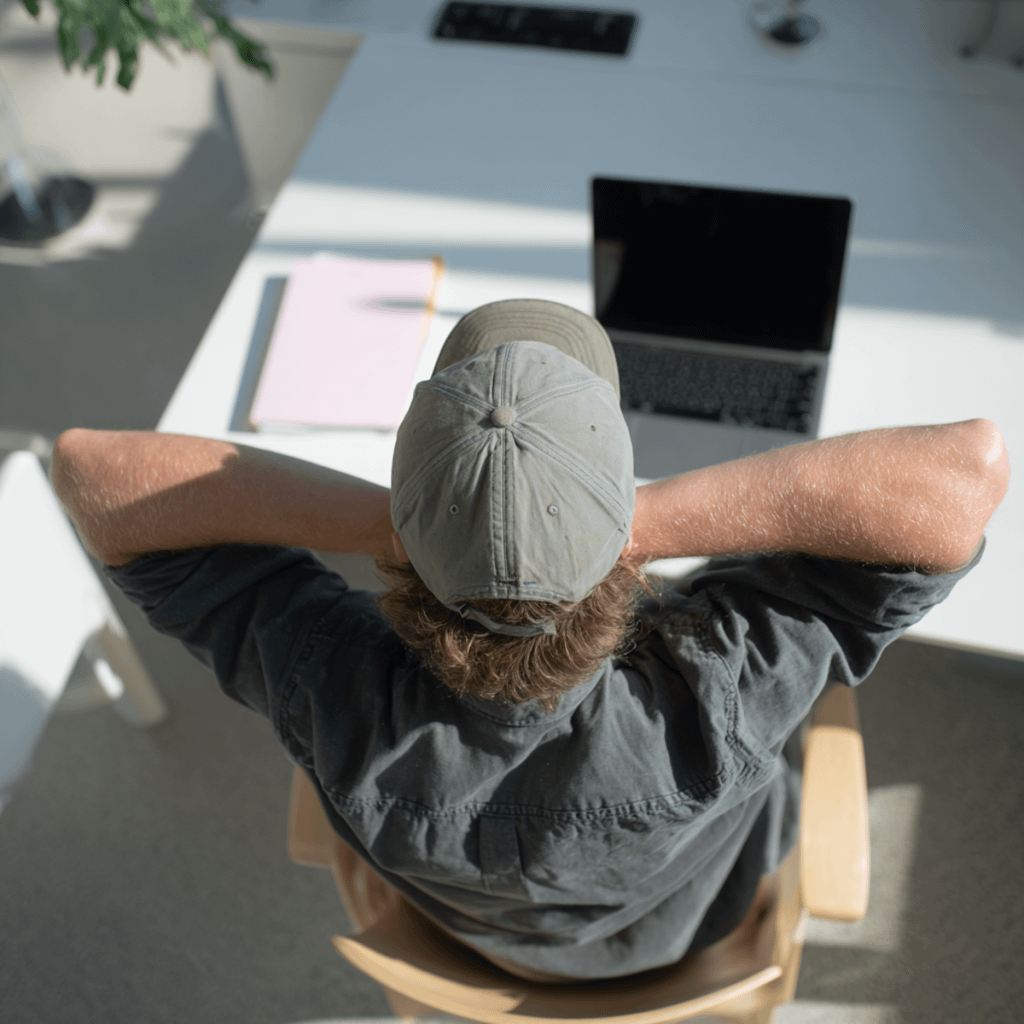 Person relaxing at a desk with a laptop and folder, viewed from behind, in a bright and modern office setting.