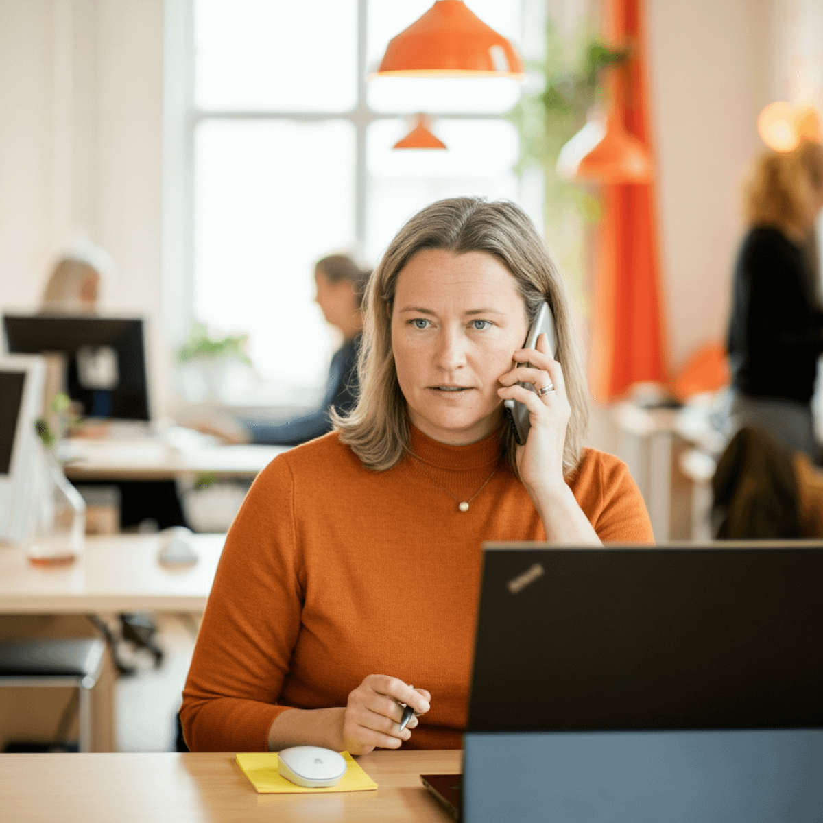 Focused woman in orange sweater on phone call at modern office desk with laptop and orange decor.