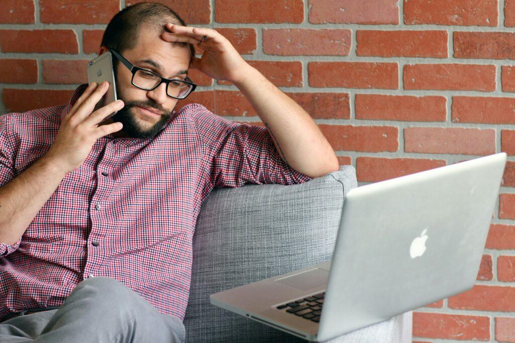 Man sitting on a couch looking stressed while talking on the phone and using a laptop, with hand on his forehead in frustration