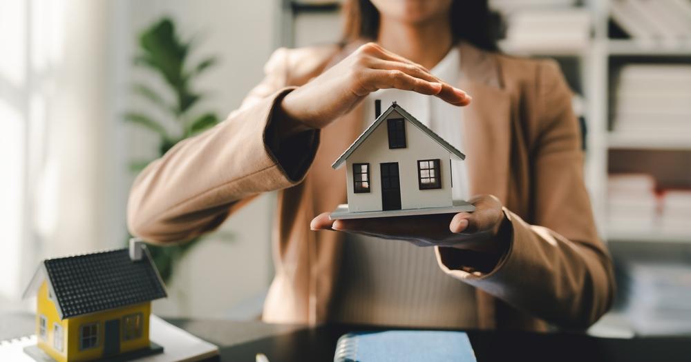 A woman holds a small house model in front of her desk, showcasing her interest in architecture or real estate.
