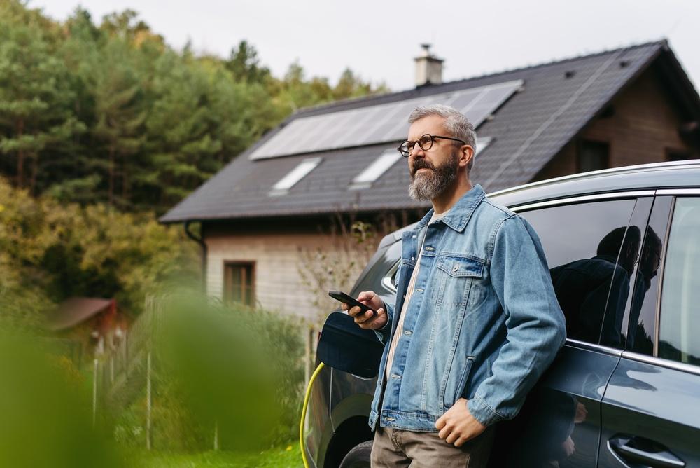 Man holding a phone leaning against a car in front of a house with solar panels