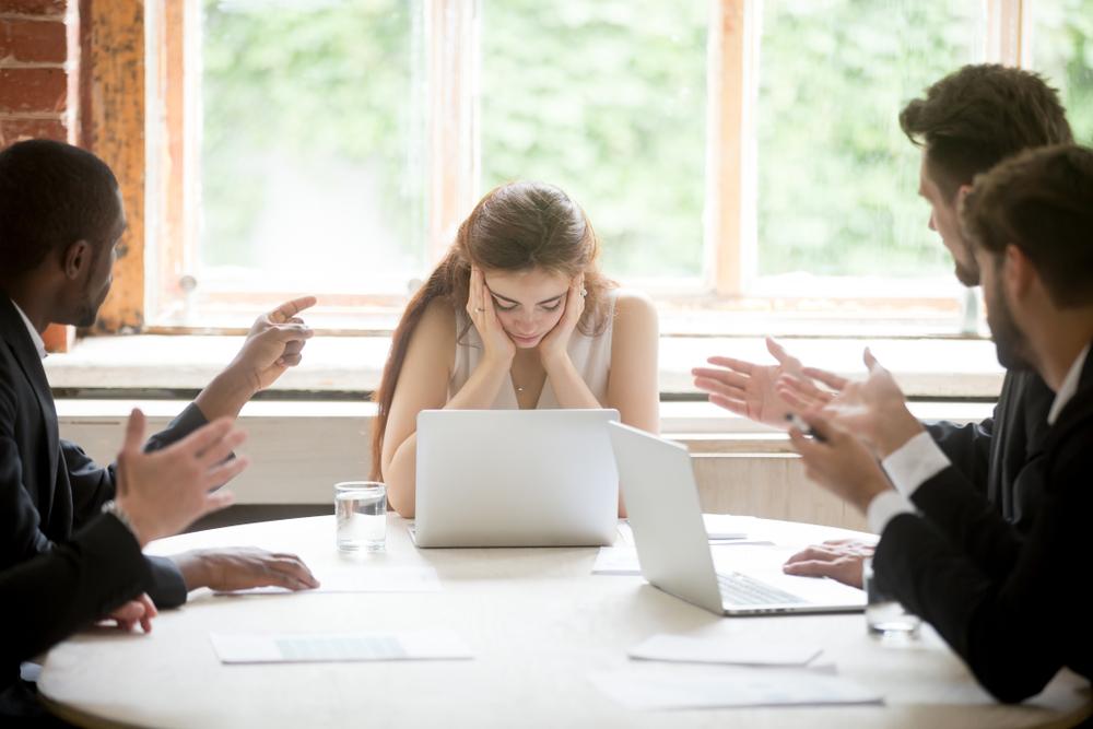 Woman holding her head between her hands at a table while several men gesture at her