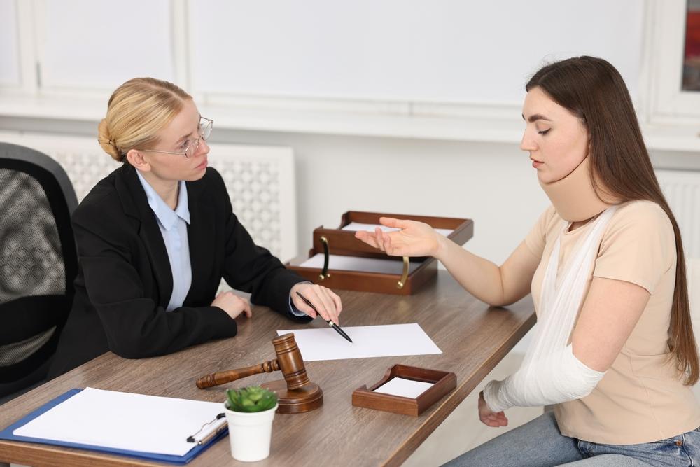 Woman with her arm in a sling and cast talking with a lawyer across a desk with a gavel and documents