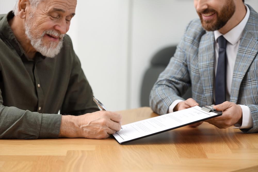 Older man signing a document on a clipboard held by a man in a suit